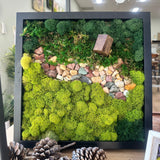 Framed moss wall art with decorative stones and pinecones on a wooden surface.