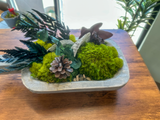 Decorative arrangement with moss, ferns, mushrooms, eucalyptus, and pinecone in a carved wood  bowl and stones